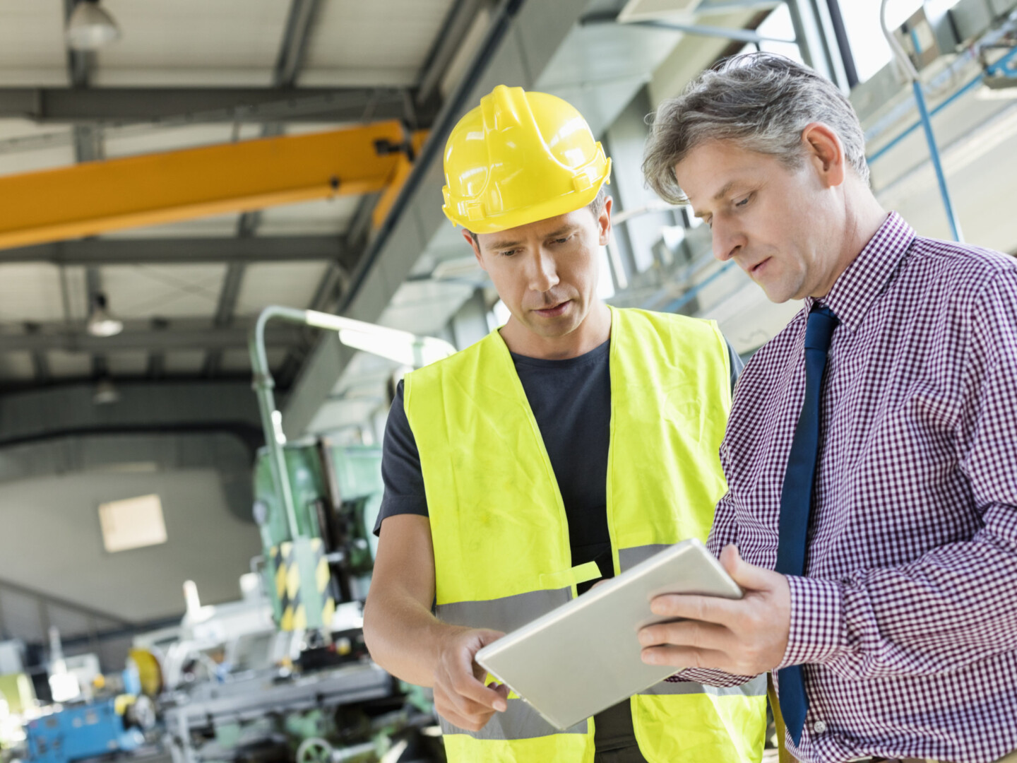 The Benefits of Outsourcing Health and Safety displayed by a worker and manager in a warehouse discussing plans over a tablet. The worker is wearing high visibility PPE and a hard hat.