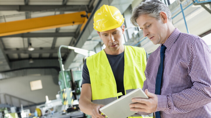 The Benefits of Outsourcing Health and Safety displayed by a worker and manager in a warehouse discussing plans over a tablet. The worker is wearing high visibility PPE and a hard hat.