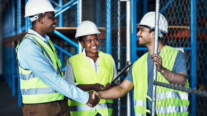 Impact of social values on business represented by three factory workers, two of which are shaking hands. They are dressed in high visibility vests and white hard hats.