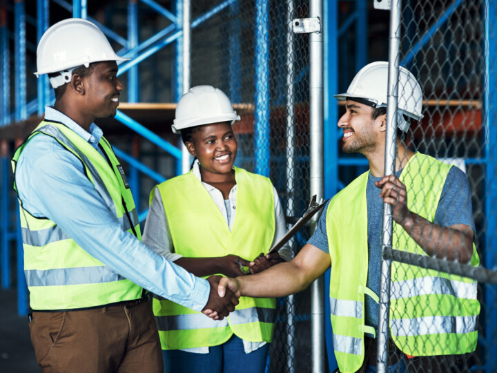 Impact of social values on business represented by three factory workers, two of which are shaking hands. They are dressed in high visibility vests and white hard hats.